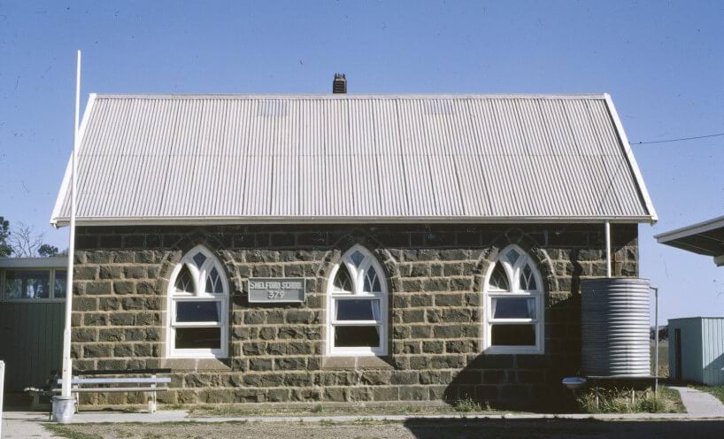 Photograph of the Shelford school building. The building is bluestone and has a corregated iron roof. It has a flag pole and a water tank. 