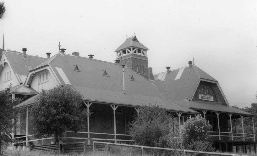 Black and white photograph of the Wandiligong school building with trees in front of the school. The school building is brick and has a bell tower. 