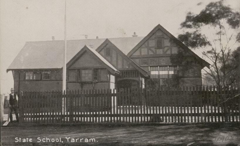 Black and white photograph of the Yarram school building. The building is brick and there is a picket fence and flagpole in front of the building. There is a man in a suit walking through the school gate.