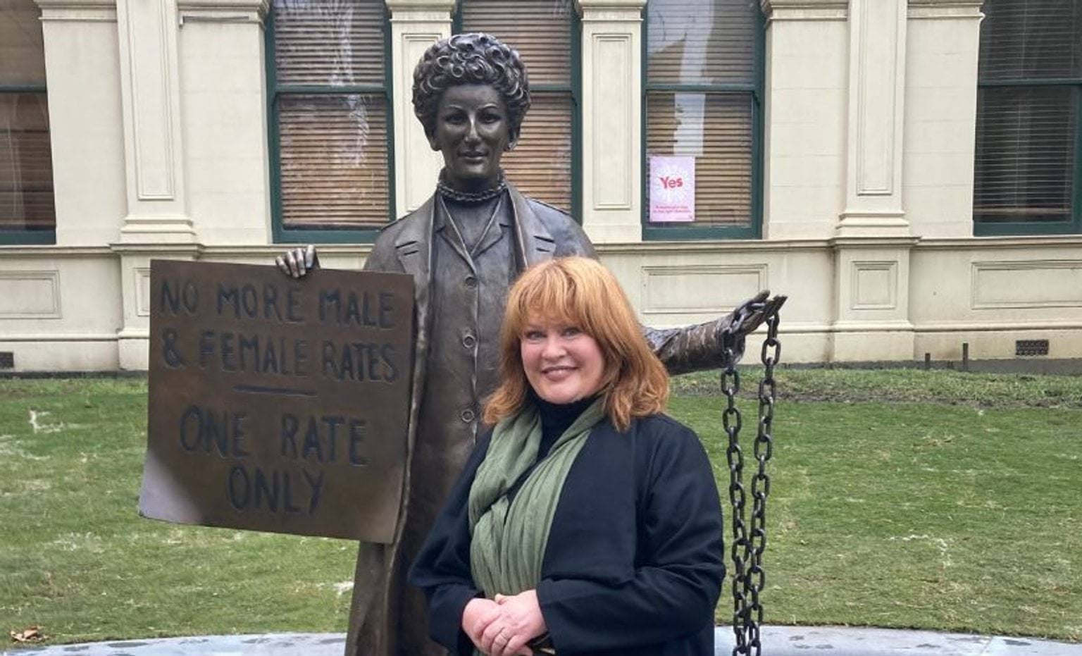 Sculptor Jennifer Mann standing with the statue of Zelda D'Aprano standing in front of the Victorian Trades Hall. She is holding a sign in her right hand that says "No more male and female rates. One rate only". In her left hand is the chain the from around her waist and would be chaining her to the courthouse.