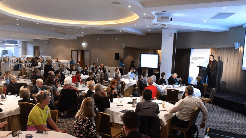 Image of attendees in a large event room listening to a speaker on stage with a microphone and screen