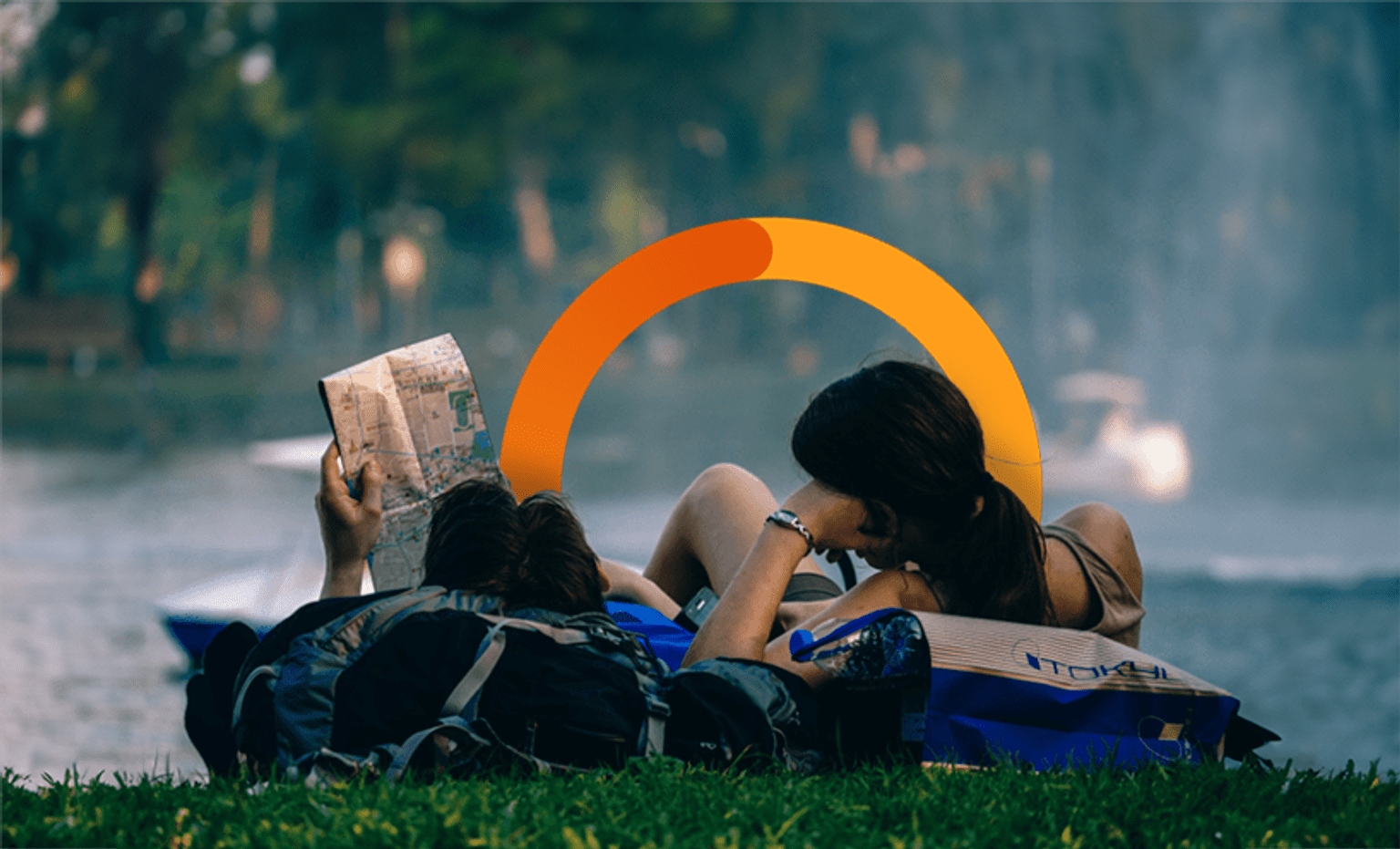 A couple are lying on the grass in front of water, looking at a map
