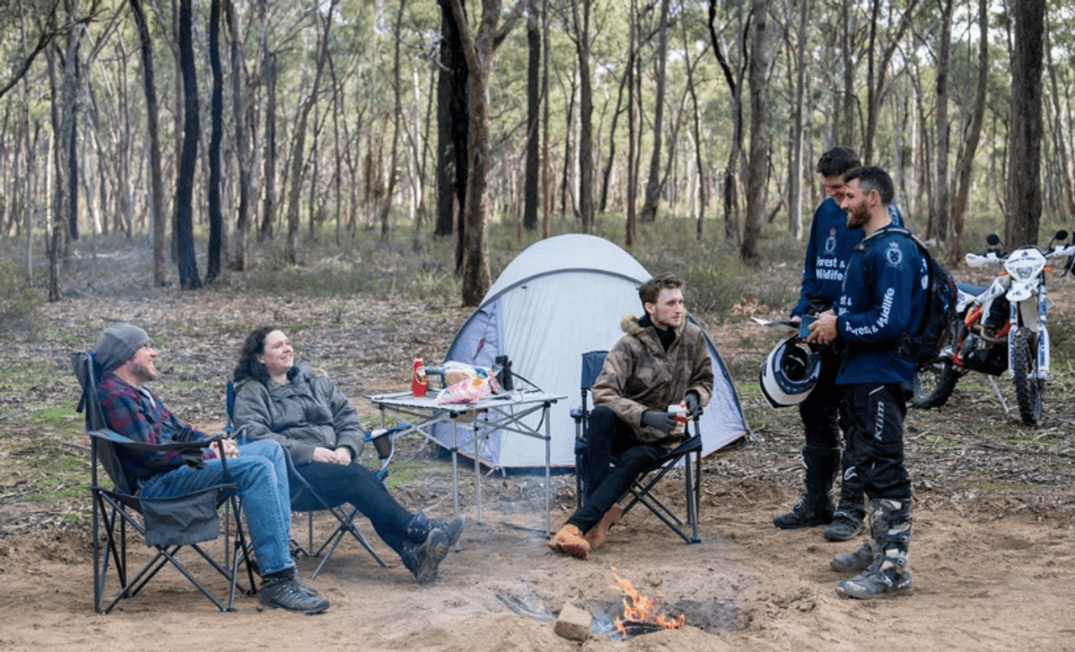 Three people sit in a forest clearing on chairs around a campfire in a small trench. They are chatting with two men in navy blue Conservation Regulator uniforms.