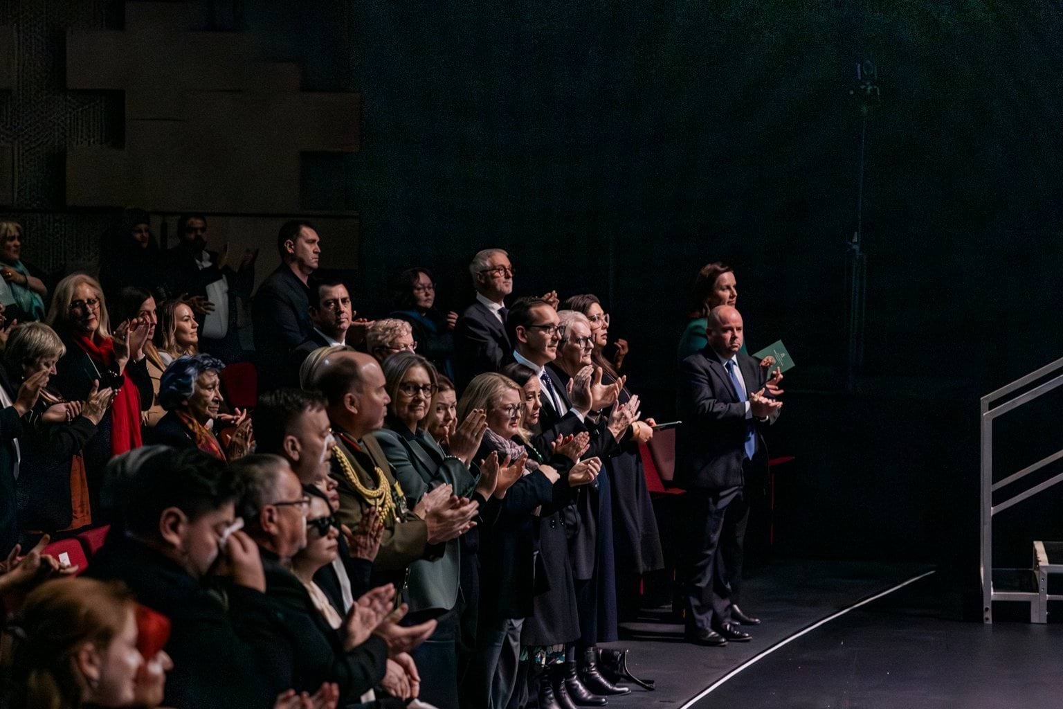 People in the audience of a dark theatre stand and clap. 