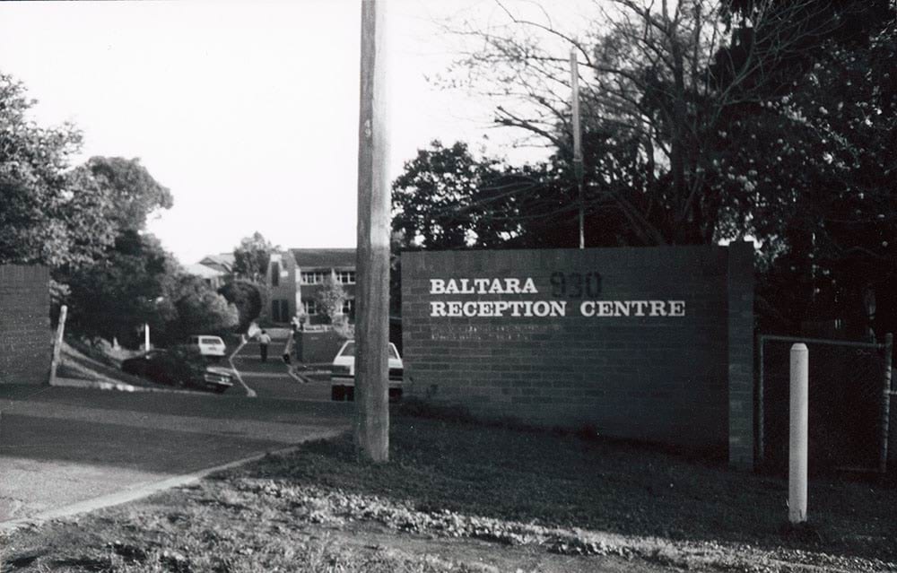 A black and white photograph of the entrance the Baltara Reception Centre. There is a brick fence on each side of the entry way. Staff have parked the cars and are walking towards the front door.