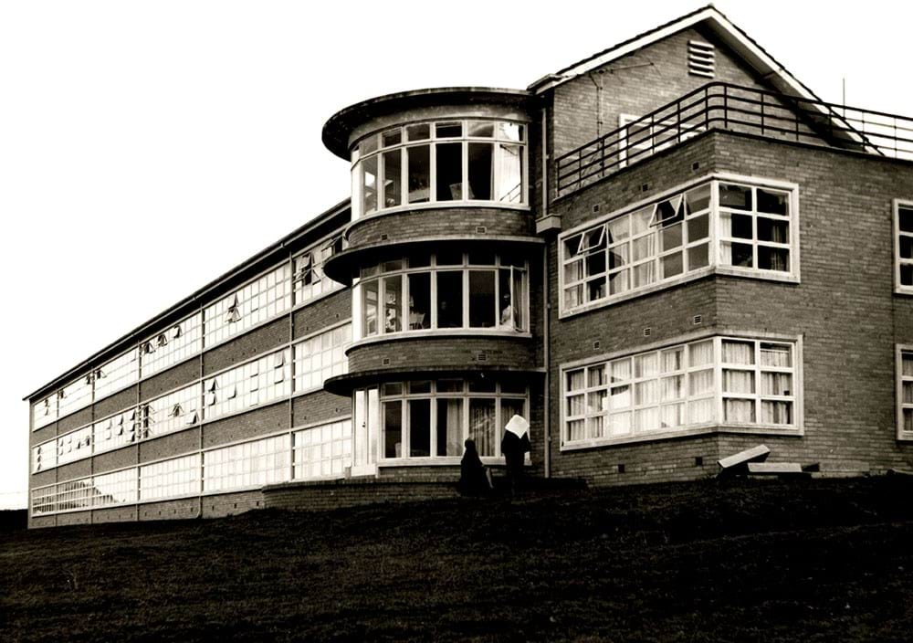 Sepia toned photo of the brick Sunbury building with large bay windows. There are nurses wearing wimples walking out front. Photo taken in 1964.