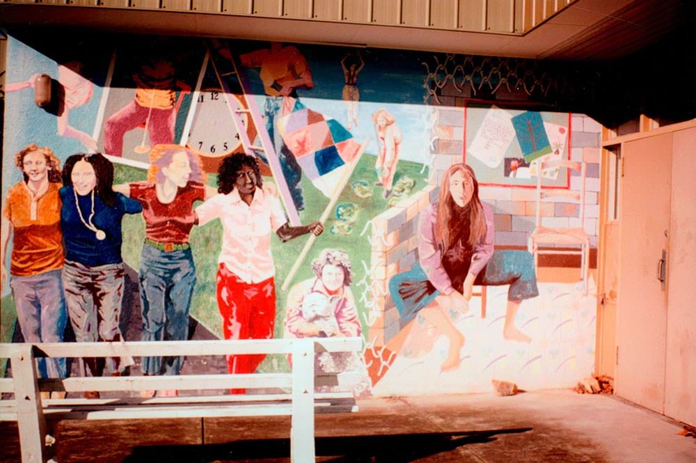 A mural on an outside wall at Winlaton Training Centre. The mural includes imagery of four young women in a row with their arms on each other shoulders and the fourth young woman is holding a flag that is a patchwork of different colours. There is a young person on a ladder, in front of a clock with no hands. A person is parachuting in behind someone else in the garden weeding cabbages. In the foreground is a young person hugging a white sheep. And there is a young woman sitting on a stool in a room.