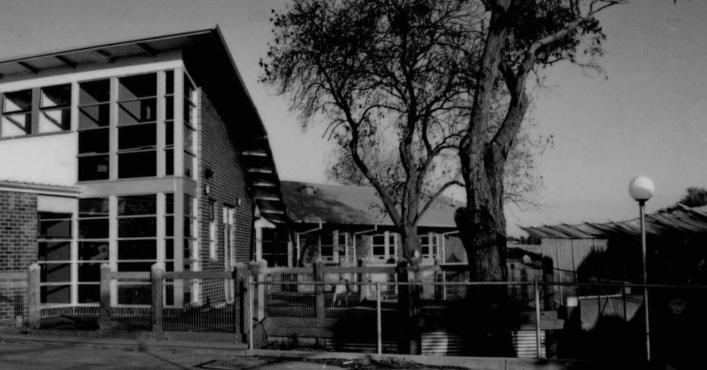 A black and white photo of the Parkville Youth Residential Centre, 1999. A brick building with a metal fence around it and two large trees in the yard.