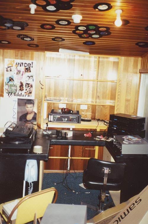 Photo of the radio room. There is acoustic wood panelling cladding the walls and the ceiling. There are small vinyl records stuck to the ceiling in flower patterns for decoration. The built in desks and shelving have radio show producing equipment, vinyl record player, cassettes, microphones and headphones.