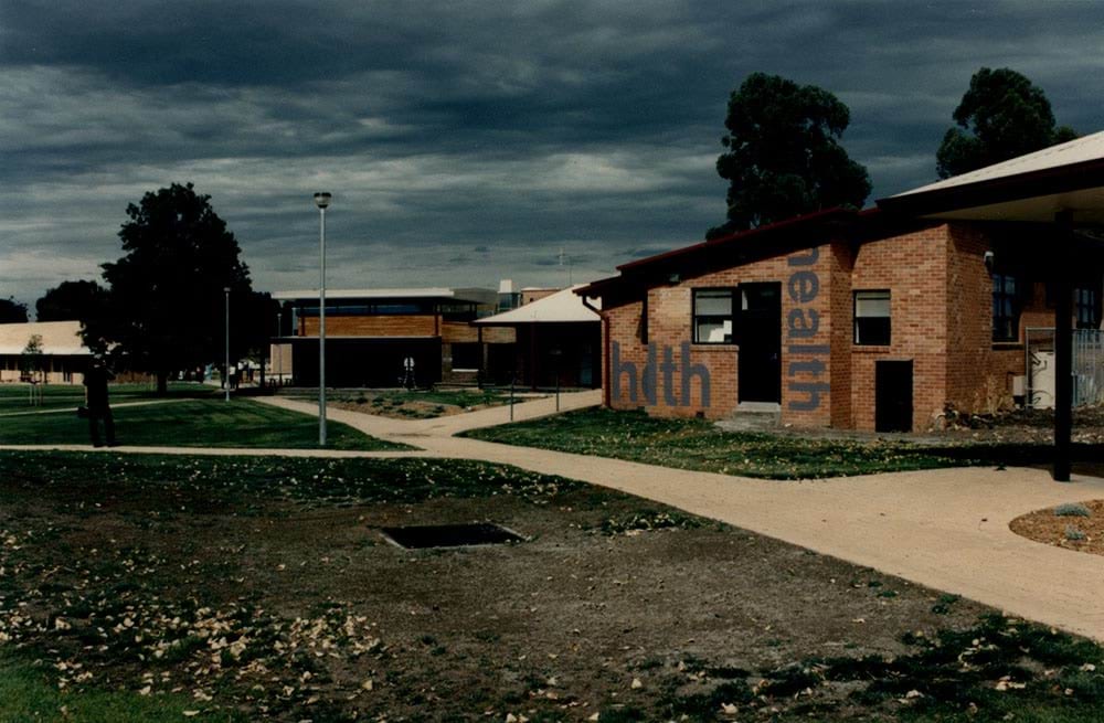 Photo of the Health building at Malmsbury Juvenile Justice Centre circa 1997. It's a cloudy day, threatening rain. There is grassy lawn on either side of the path to the Health building and the other buildings in the background.