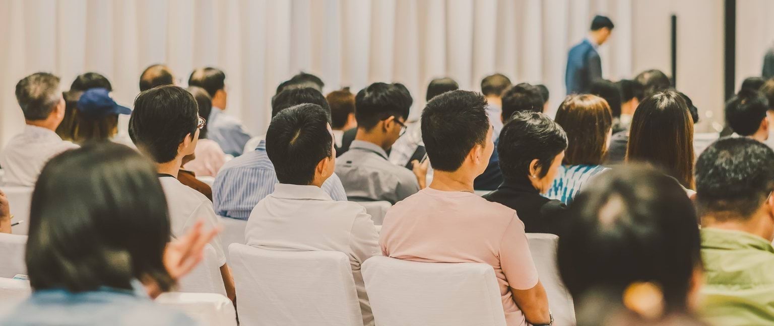People seated in a row of seats inside a community hall. 