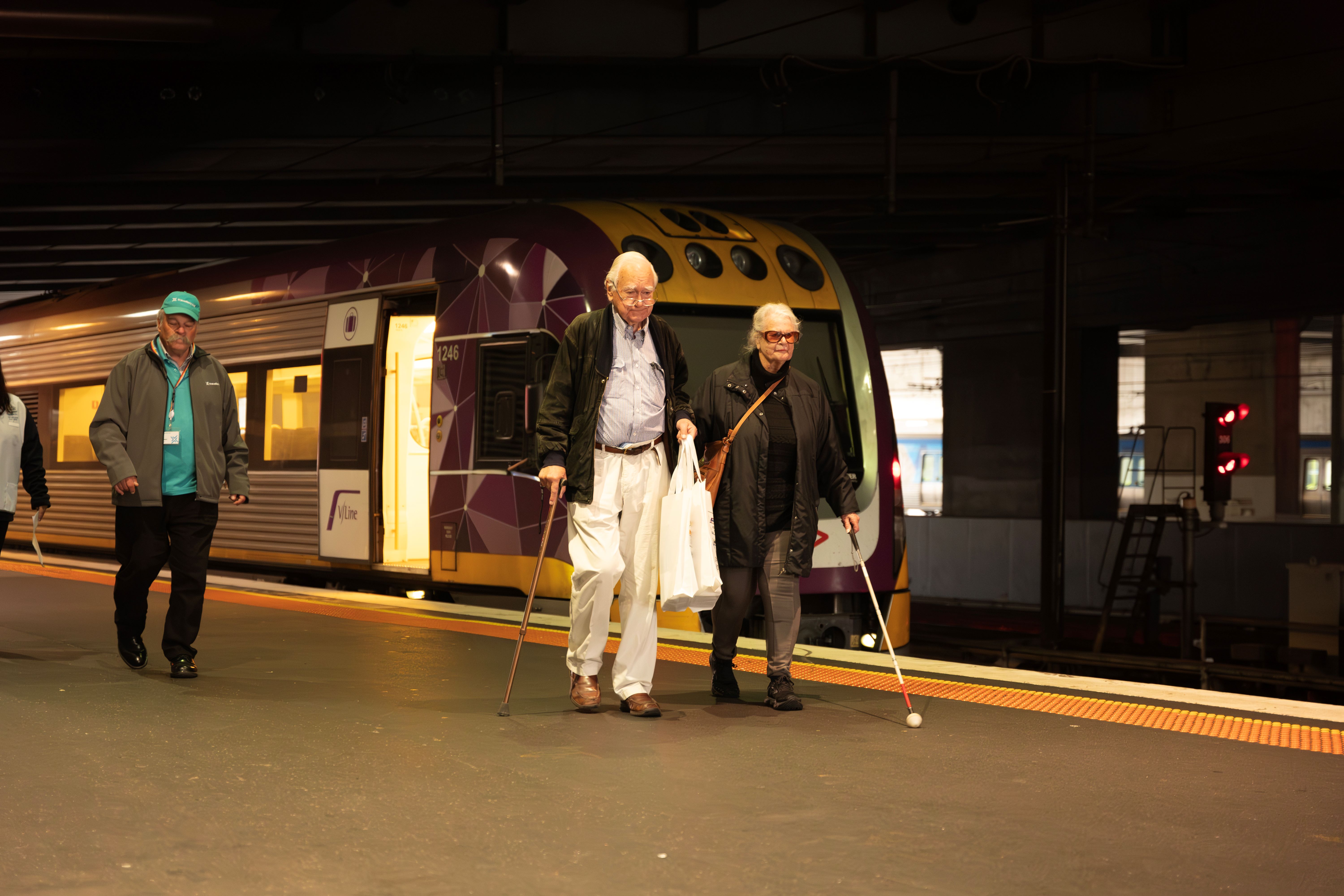 Two people walking on a platform at Southern Cross Station at try Before You Ride. One person is a white cane user and the other person is using a walking stick. A V/Line train is in the background.