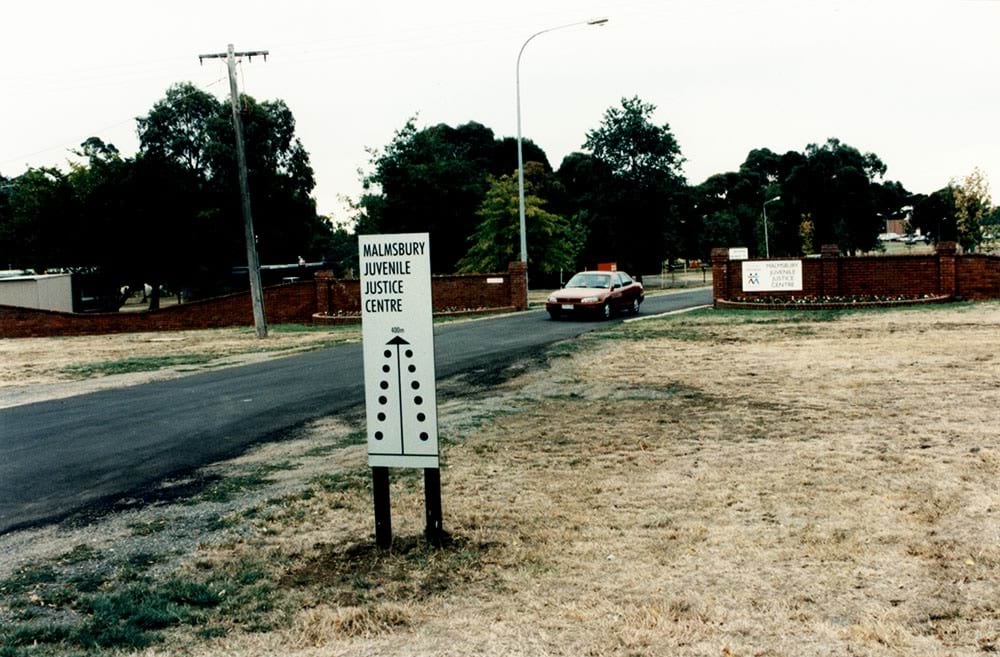 A phot of the front entrance to the Malmsbury Juvenile Justice Centre in 1990. There is a maroon sedan car driving out.
