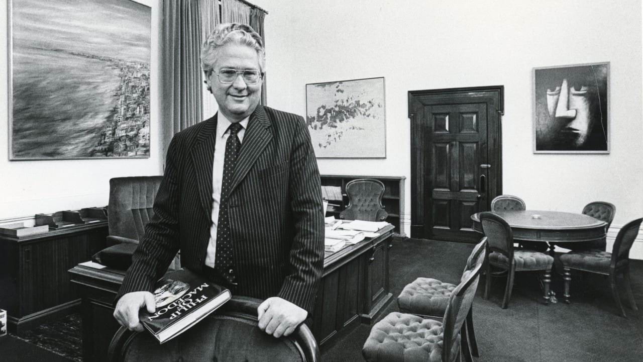 The Honourable Dr Race Mathews is wearing a pin-stripe suit and glasses. He leans on the back of a chair in his office holding a book titled Philip of Macedon