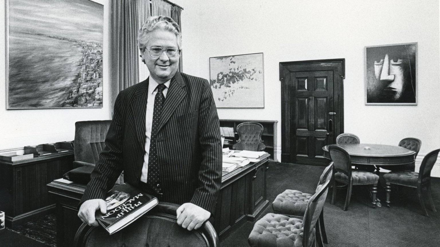 The Honourable Dr Race Mathews is wearing a pin-stripe suit and glasses. He leans on the back of a chair in his office holding a book titled Philip of Macedon