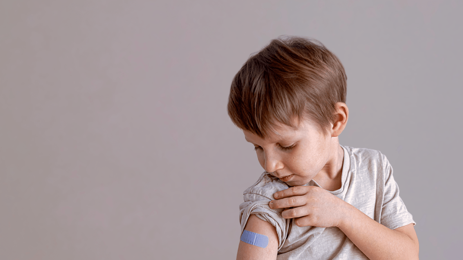 A young boy lifts up the sleeve of his t-shirt and is looking at a blue band aid placed just below his shoulder, which shows where his flu vaccination was administered