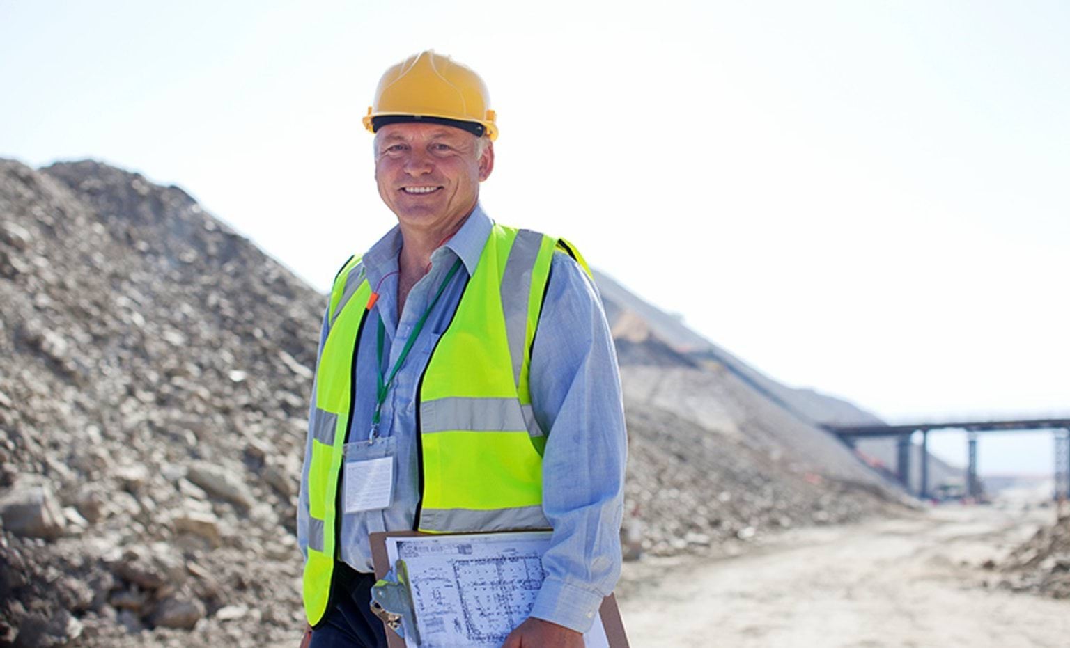 A miner standing in front of a mining site wearing a helmet and holding a clipboard