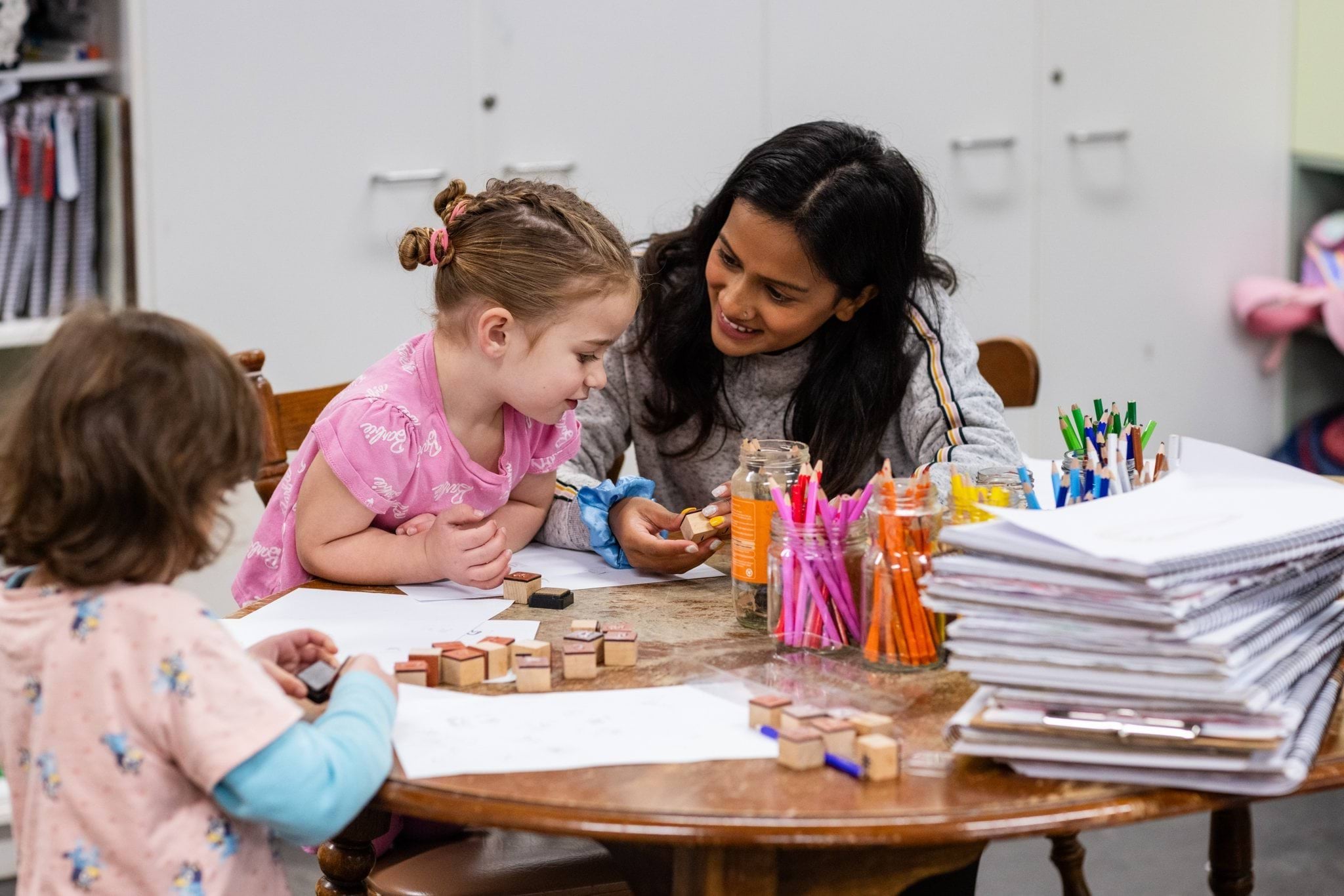 An educator supports a young girl playing with stamps and pencils at a wooden table