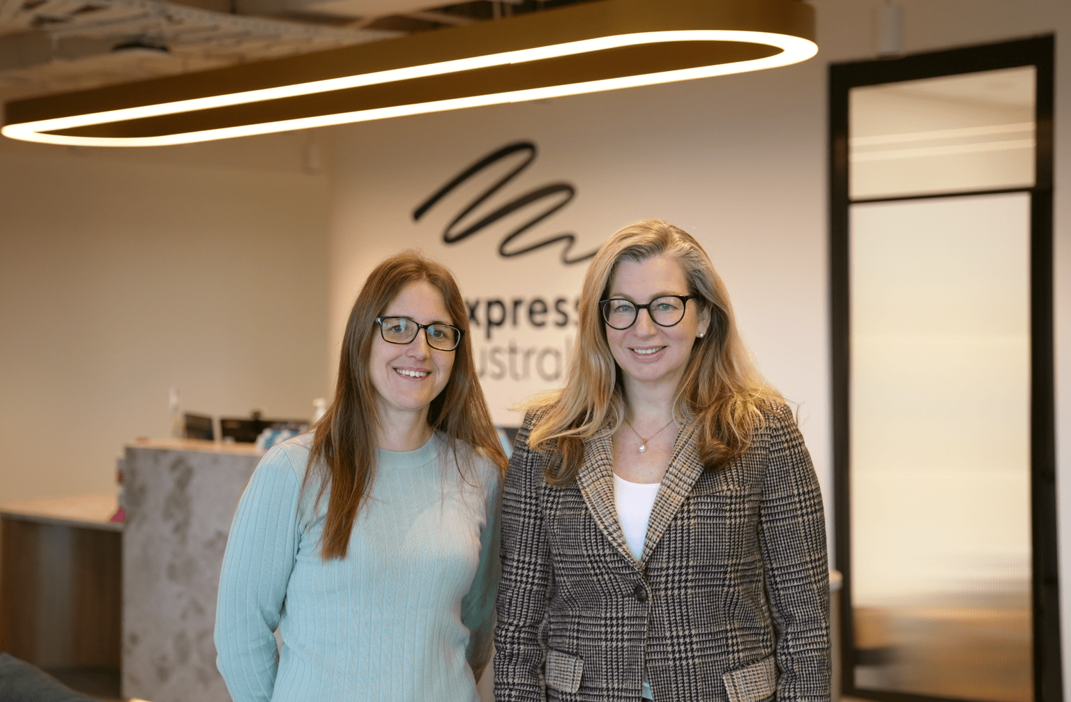 Two smiling women wearing glasses stand in the foyer of Expression Australia. 