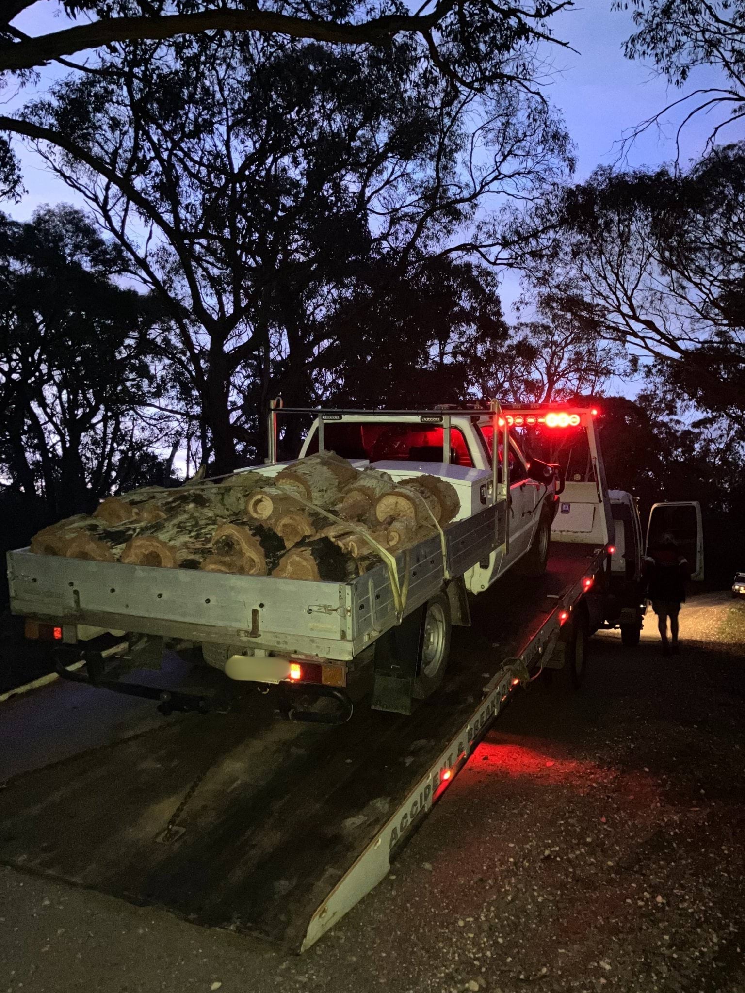 A ute loaded with firewood being loaded on to a tow truck on  a forest track