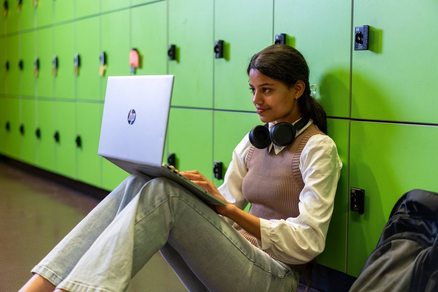 A student sitting on the floor using a laptop, resting against a row of green lockers.