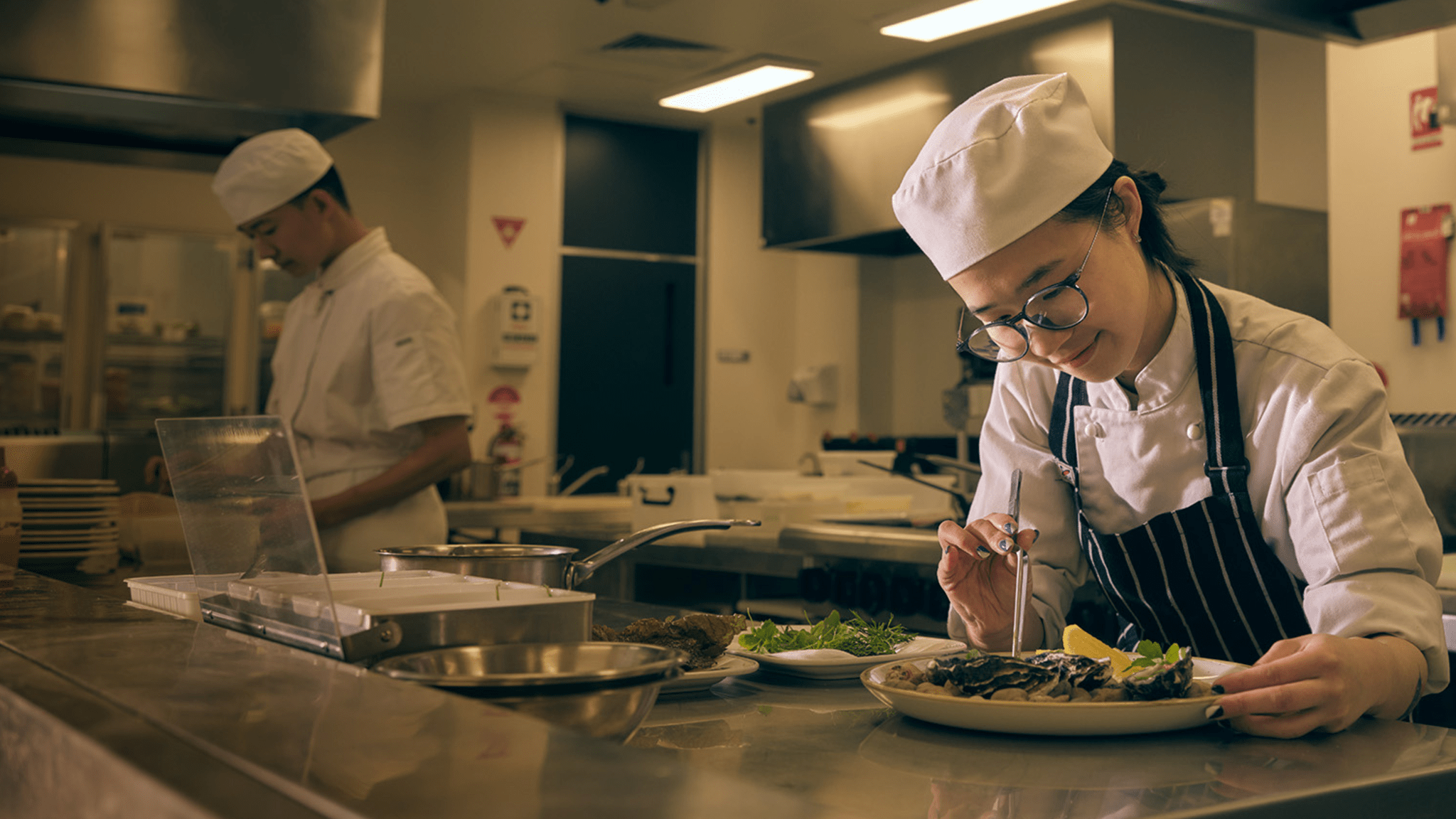 A person in a chef's uniform and apron uses tweezers to place food on a plate in a commercial kitchen.