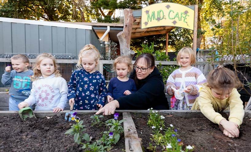 teacher and children planting flowers in a garden bed.