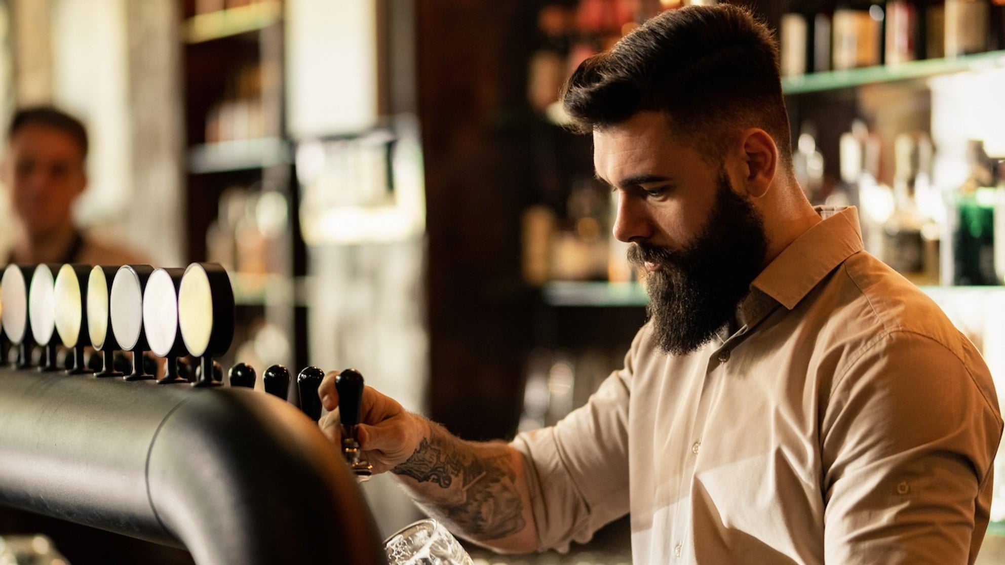 Person wearing a light-colored long-sleeve shirt pours a drink from a row of beer taps at a bar, with shelves of liquor bottles visible in the background. The person has a tattooed forearm and is holding a glass under the tap.