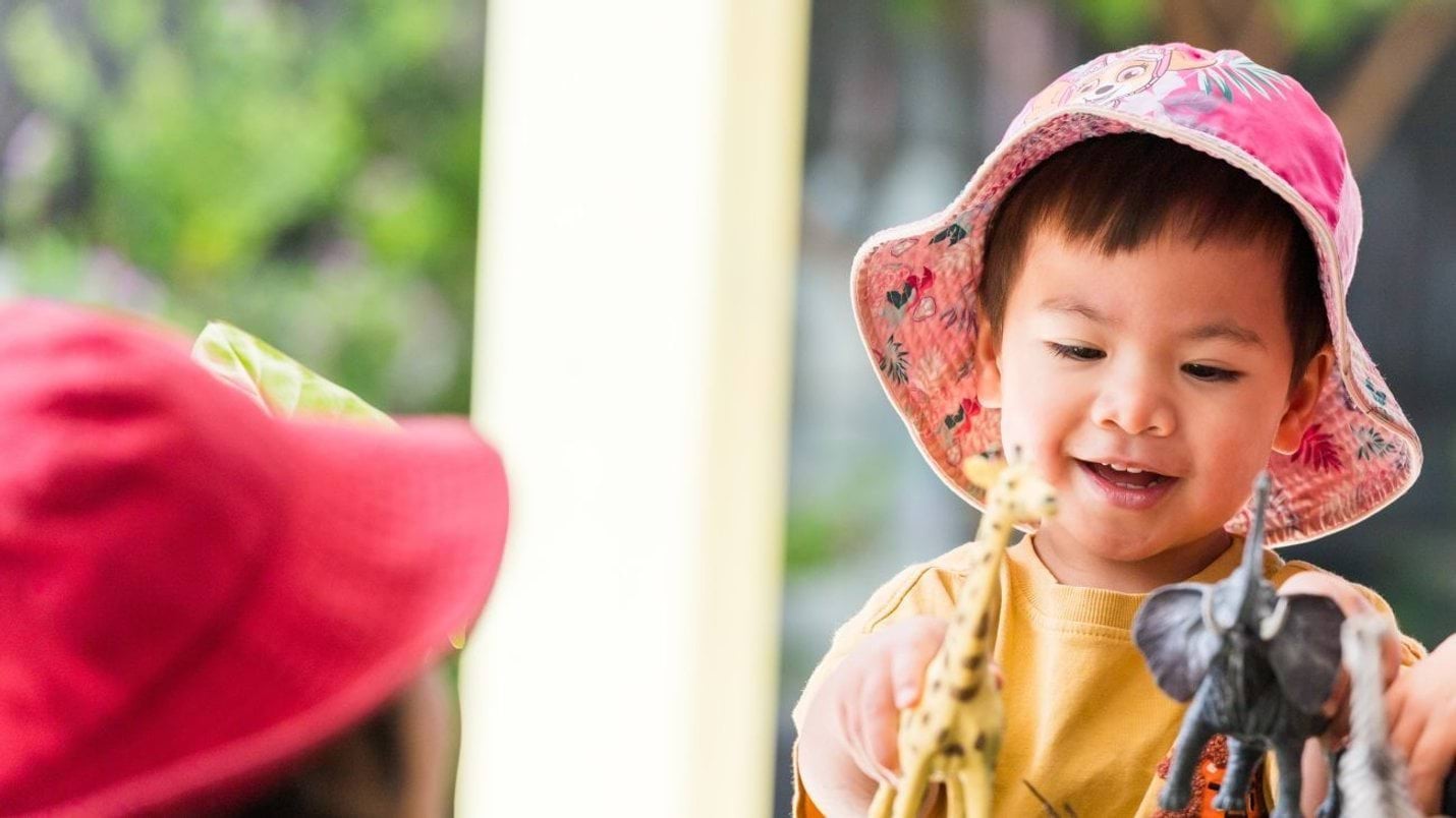 A child in a pink hat and yellow t-shirt smiling and playing with toy giraffe and elephant figurines.