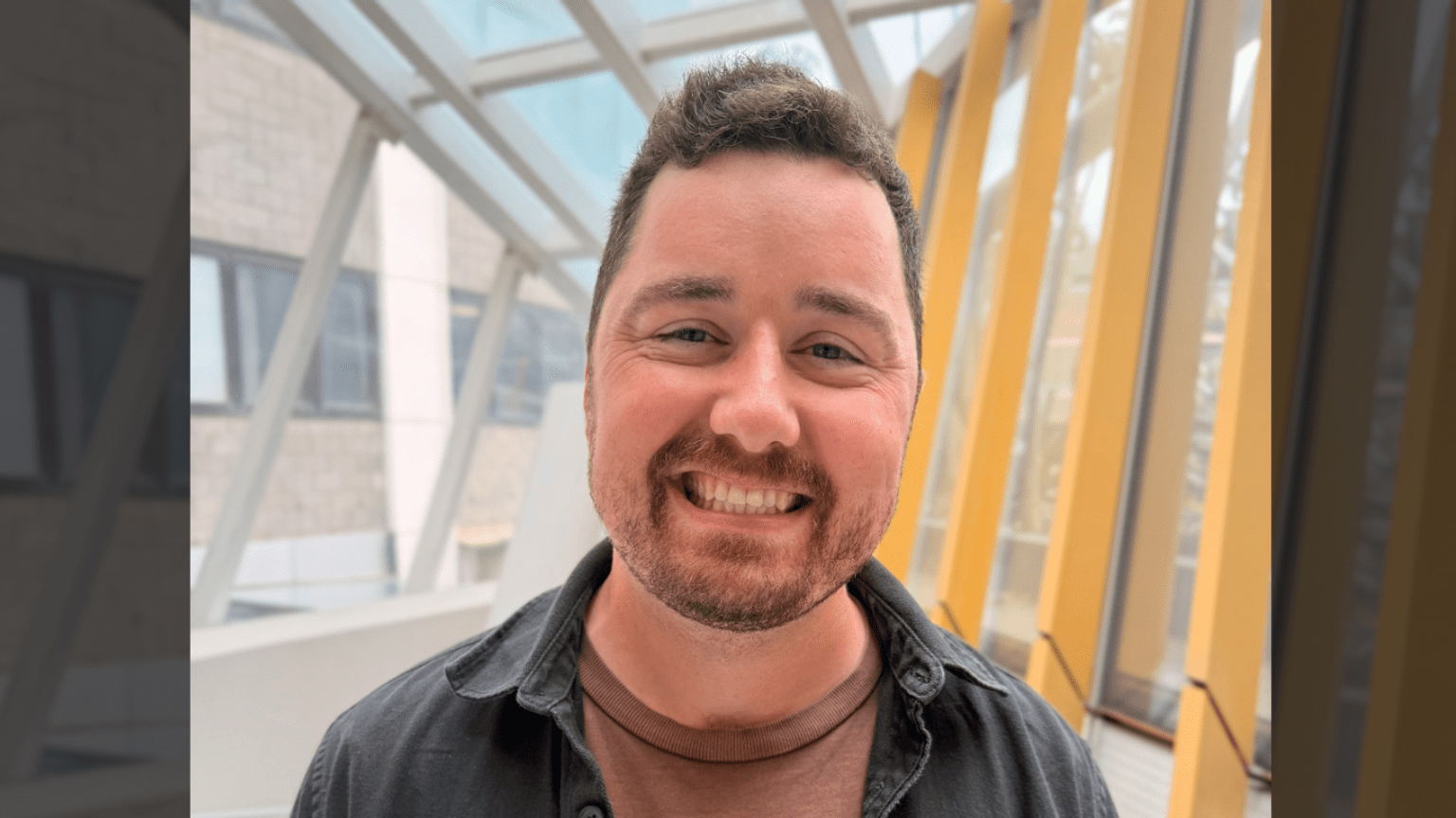 Jessy has dark hair and a beard and is smiling in a brightly lit room with large windows.