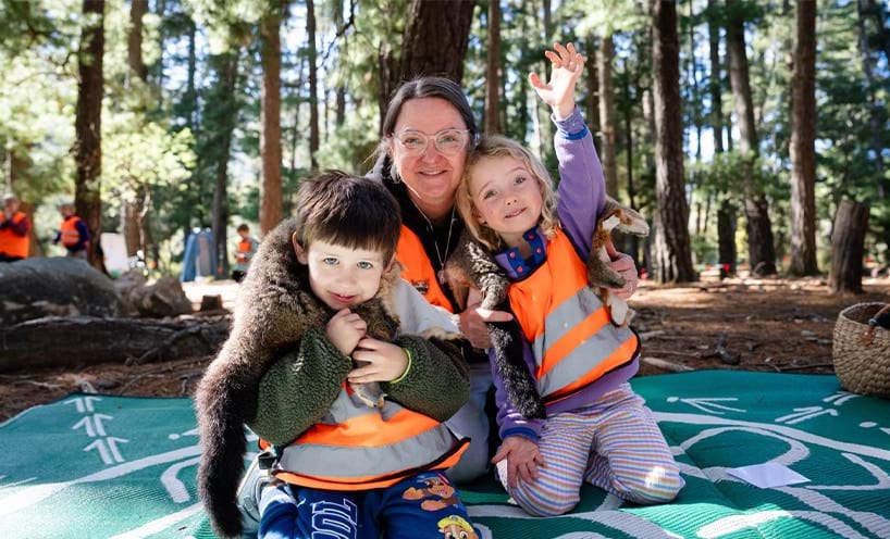 Teacher and 2 small children kneeling on a blanket in the bush.
