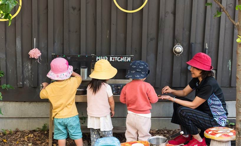 A teacher and 3 small children playing in an outdoor kitchen.