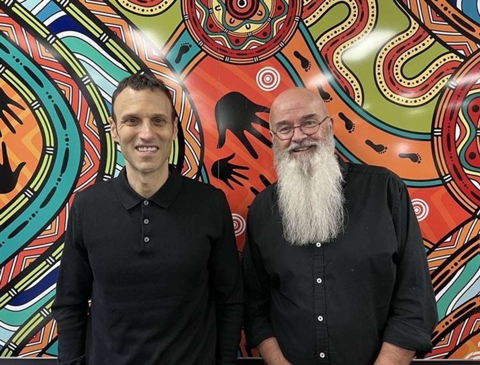 Social Services Regulator Jonathan Kaplan and Anthony Kolmus, former SSR Director, Child Safeguarding and Cultural Safety and former Human Services Regulator, pictured standing together and smiling against the backdrop of colourful First Nations artwork