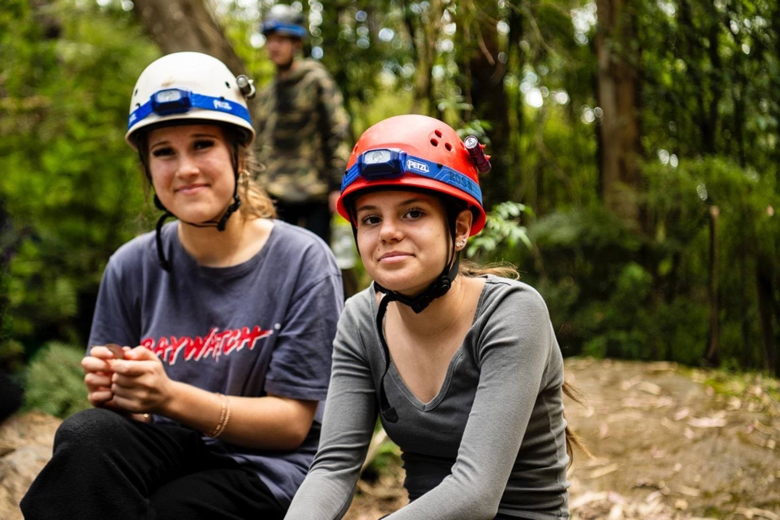 Two teenage girls sit down together in bushland.