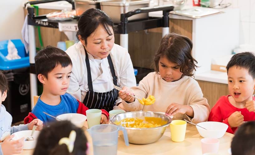 Children and a teacher sitting around a table. One child is spooning food from a bowl.