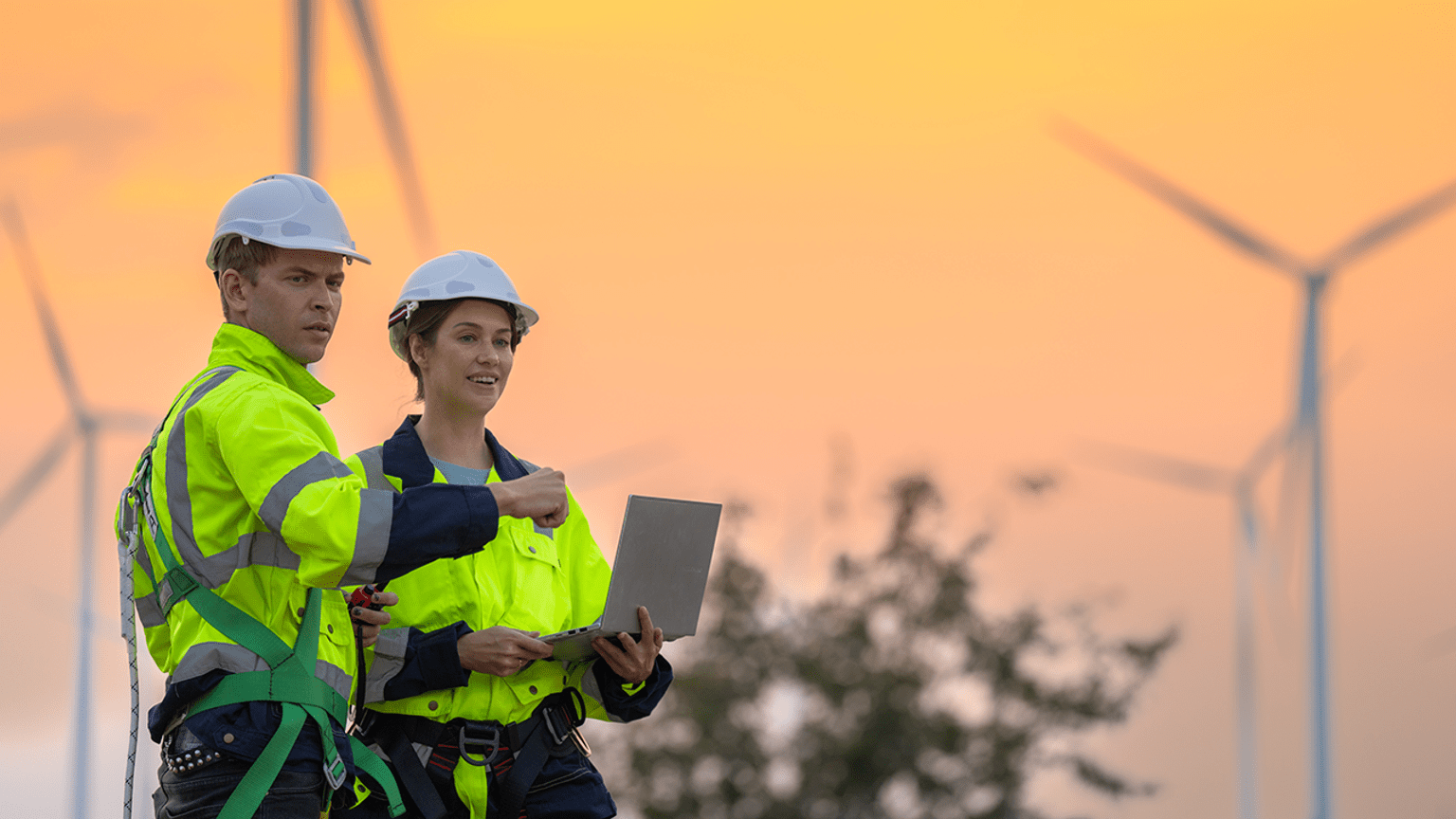 Two workers with harnesses and hard hats using a laptop with power generating windmills in the background 