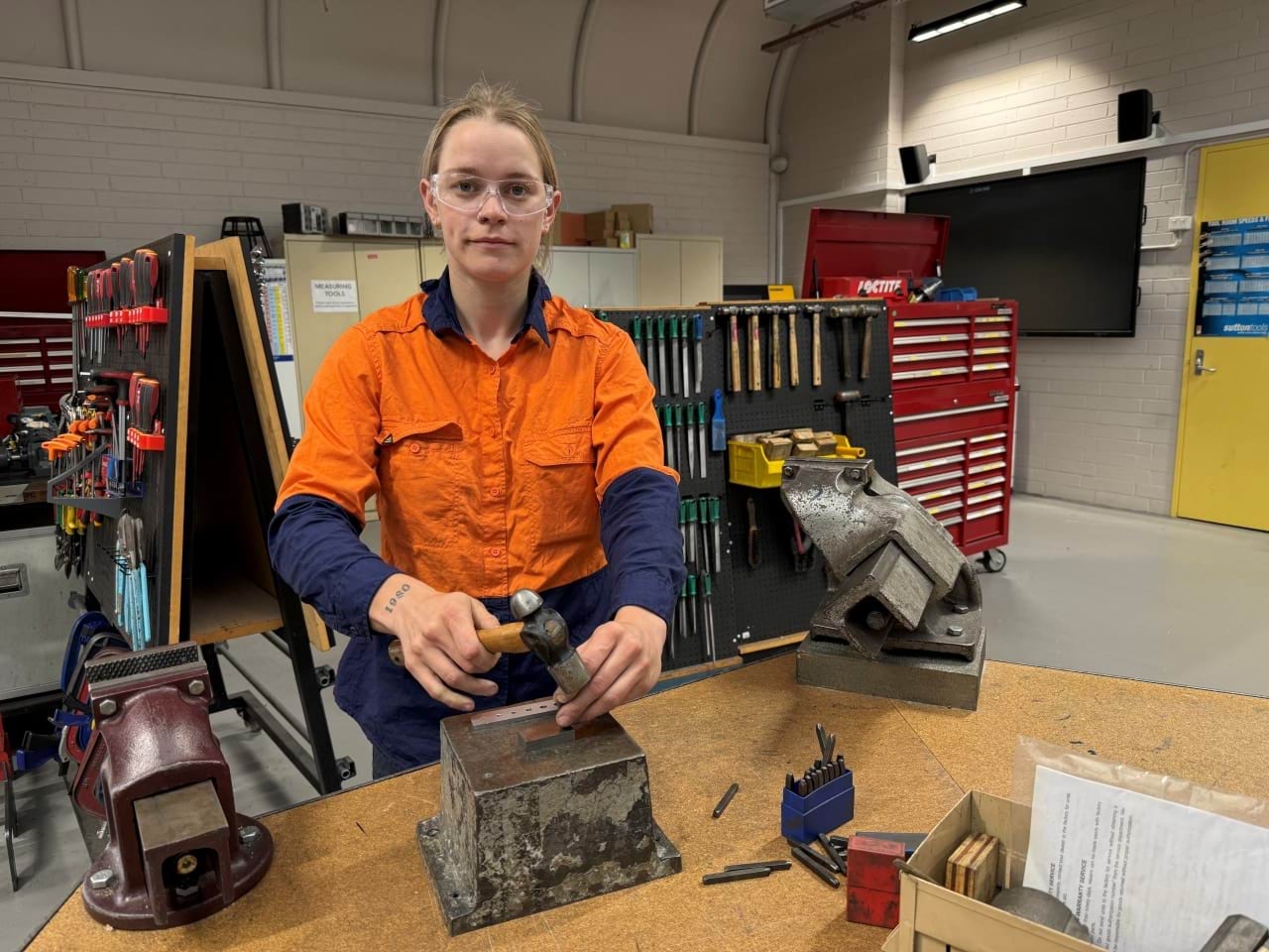 Madi is wearing a hi-vis shirt and safety glasses, using tools in a workshop.