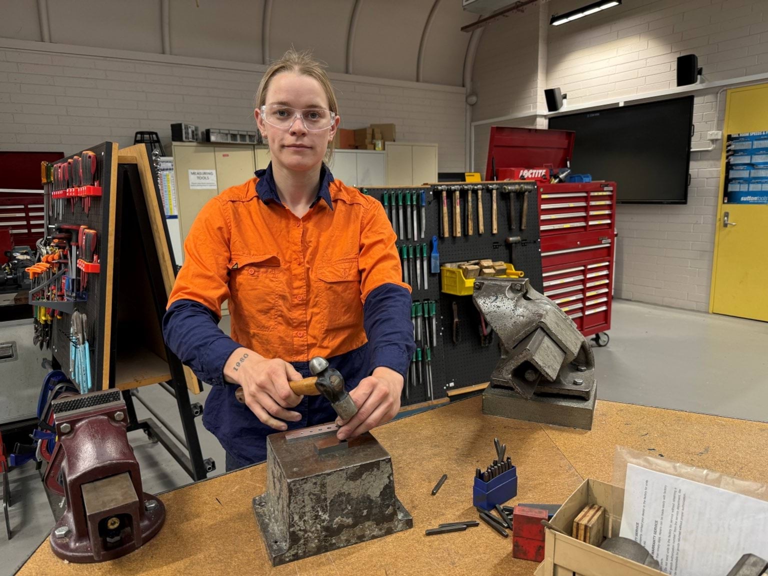 Madi is wearing a hi-vis shirt and safety glasses, using tools in a workshop.