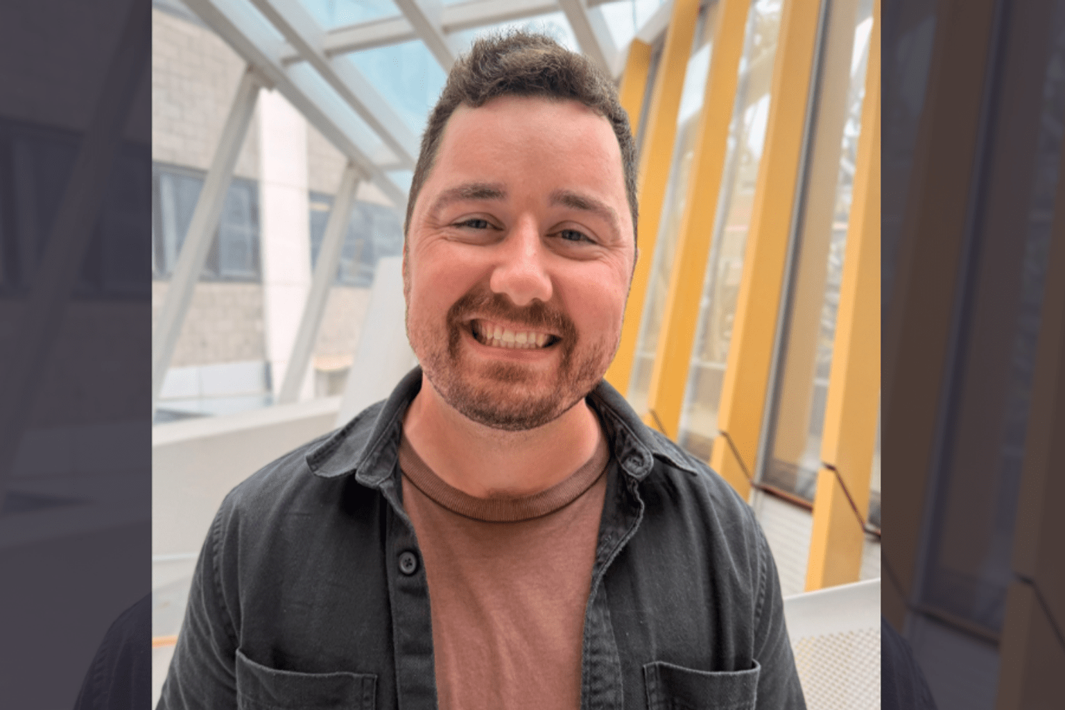 Jessy has dark hair and a beard and is smiling while standing in a bright room with large windows and beams.
