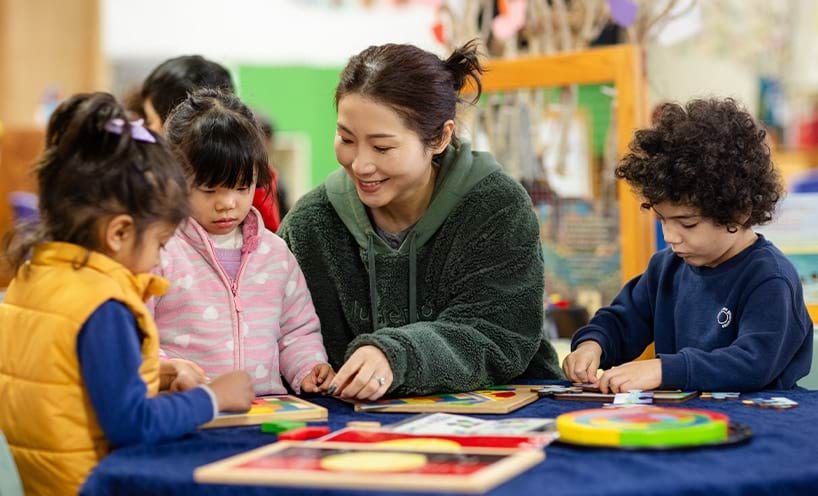 An educator assisting a group of children around a table with puzzles