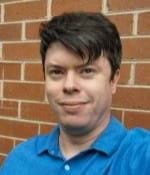 A photo of Callum Flint. He has short brown hair and is wearing a blue shirt. There is a brick wall in the background.