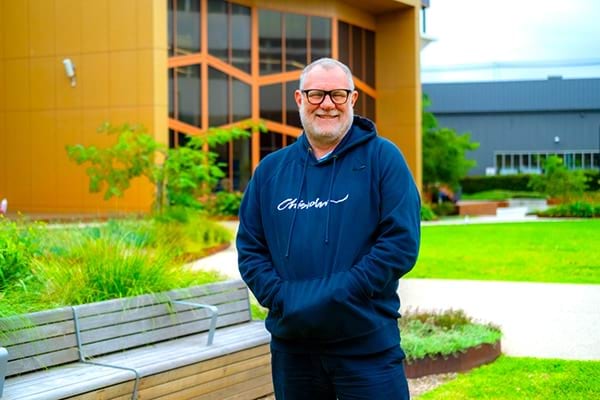 Chris, wearing a Chisholm jumper stands outdoors at campus, reflecting his journey with Free TAFE which helped him become an IT teacher. 