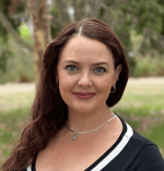 A photo of Katrina Moody in a park. She is wearing a black and white top, has long reddish brown hair and is looking at the camera.