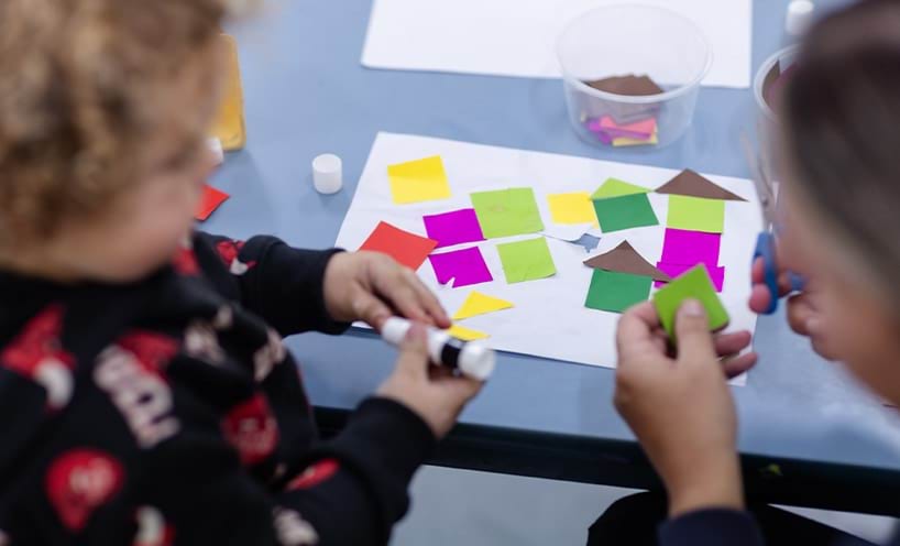 An educator assisting a child sticking coloured paper cutting on a sheet of paper using a glue stick