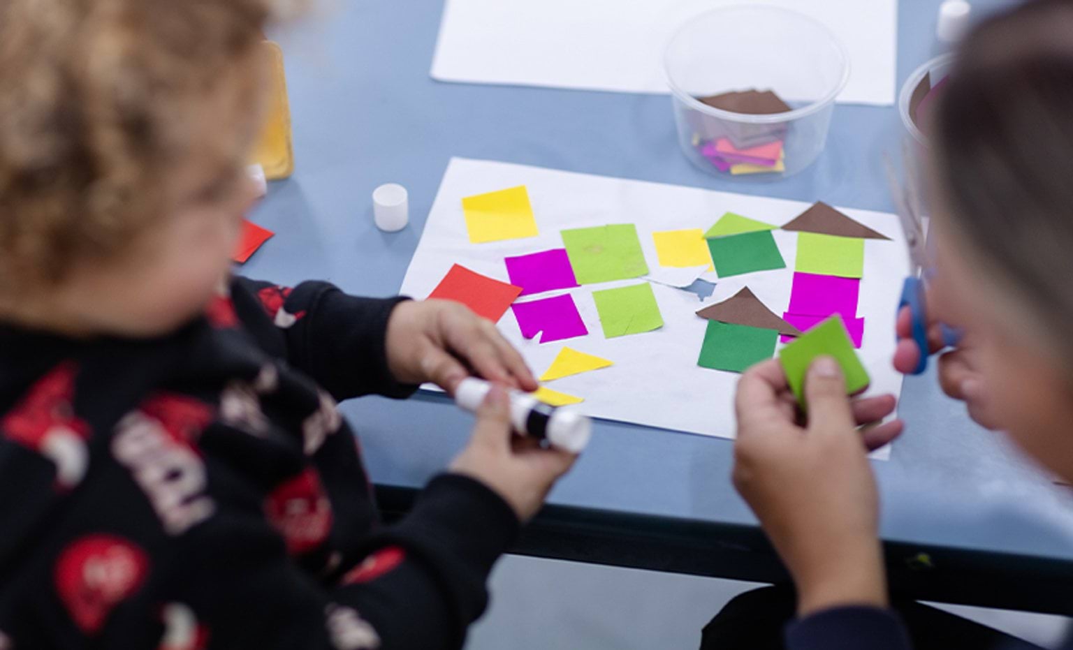An educator assisting a child sticking coloured paper cutting on a sheet of paper using a glue stick