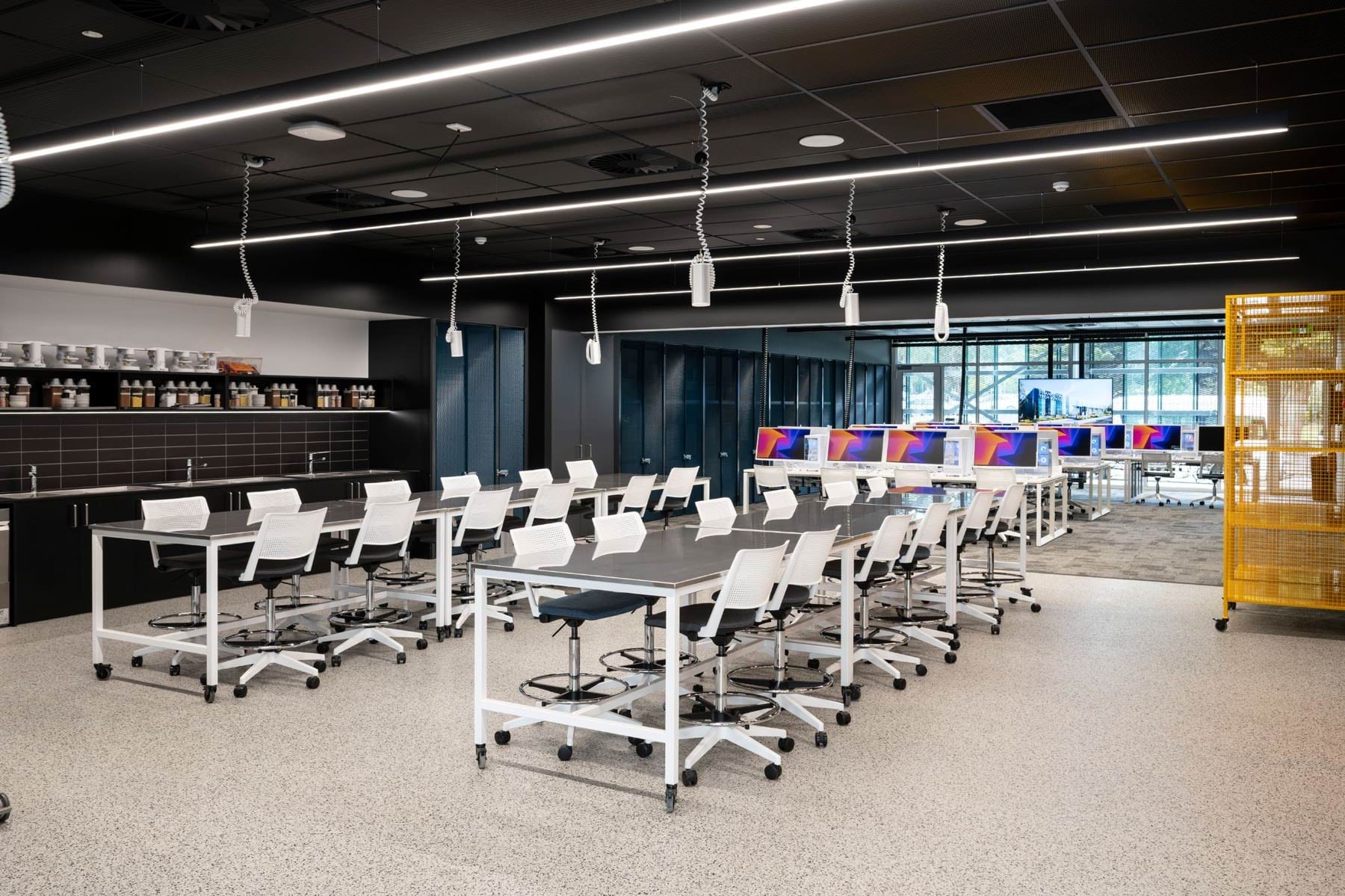 Modern classroom with long worktables, white swivel chairs, ceiling-mounted power cords, and computer stations in the background.