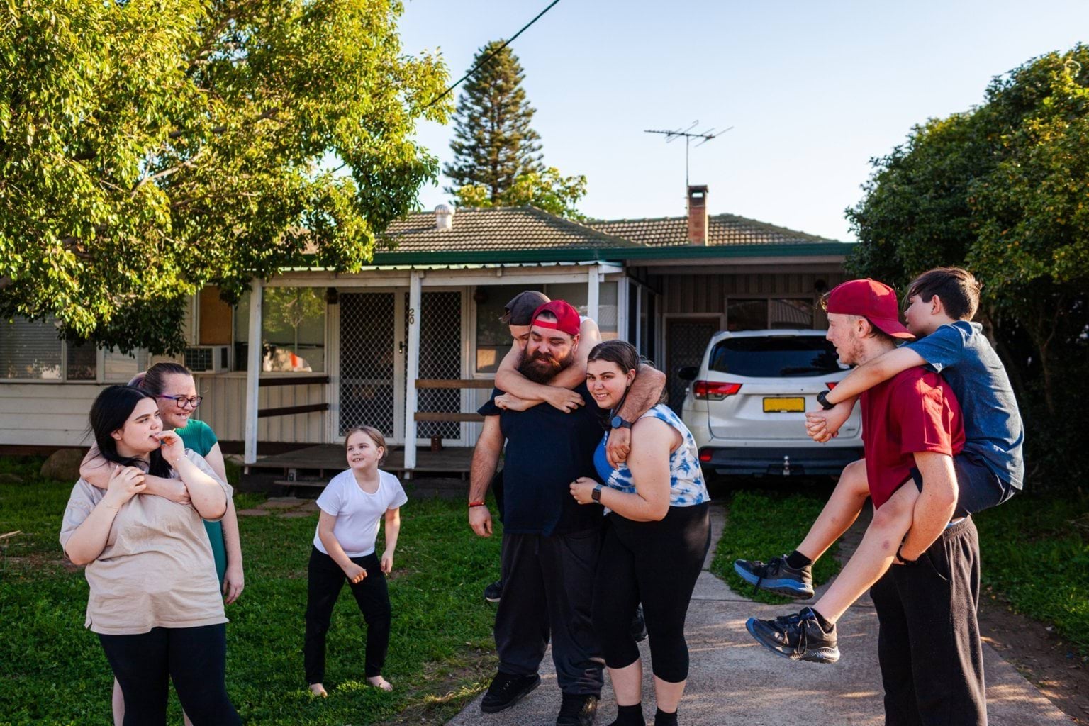 A family of 8 people hang hug and play outside the front of a house on a sunny evening.