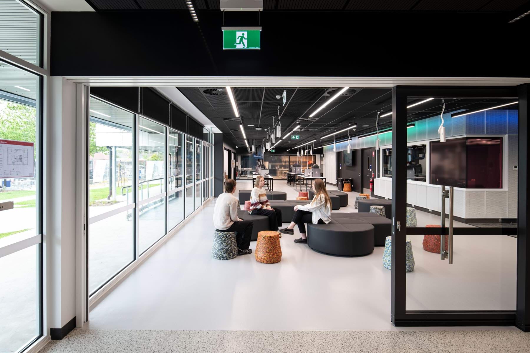 Contemporary school common area with students sitting on round seating, glass walls on one side, and workspaces in the background.