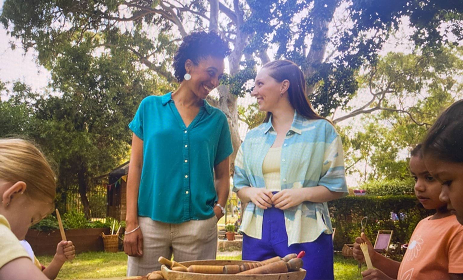 Two educators looking at each other standing next to each other and smiling. 3 children playing outdoors