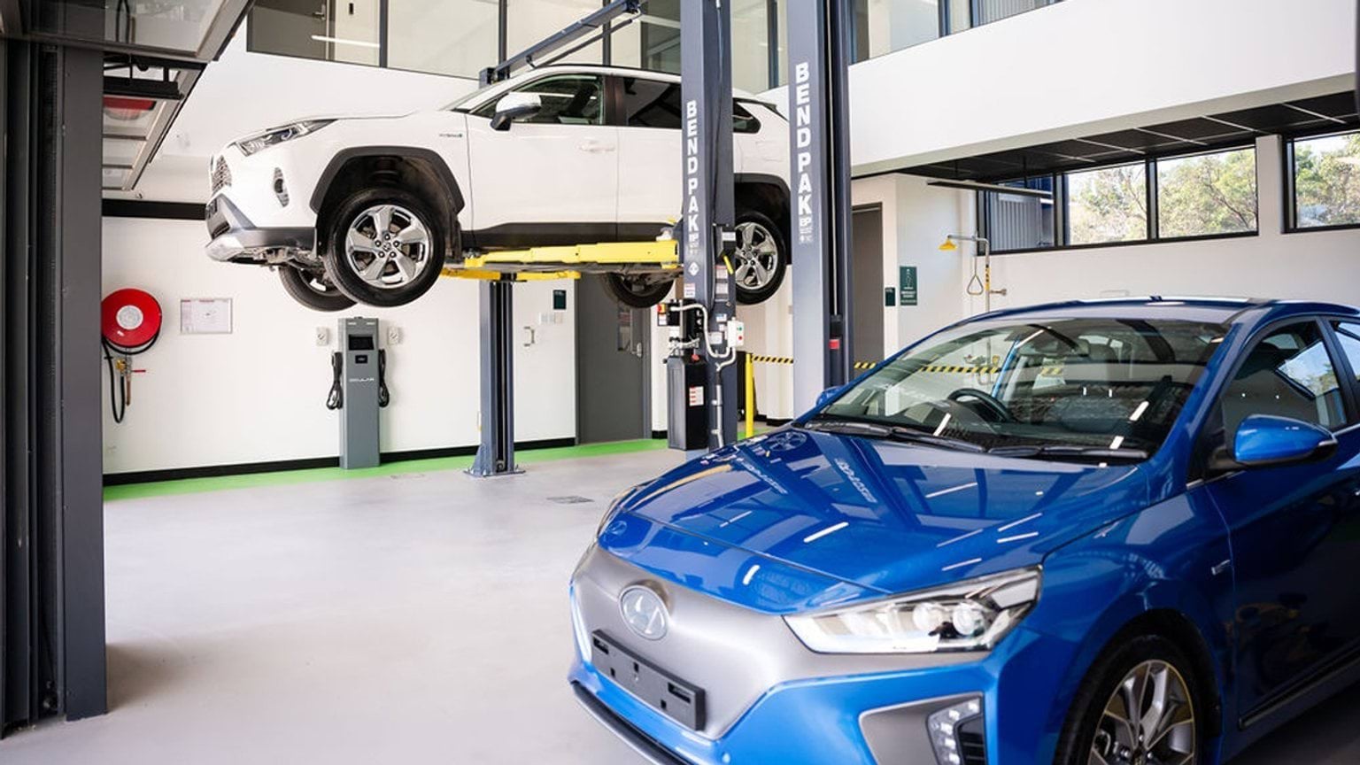 Automotive training workshop with two vehicles on hoists, including an electric car in the foreground and one car raised for servicing.